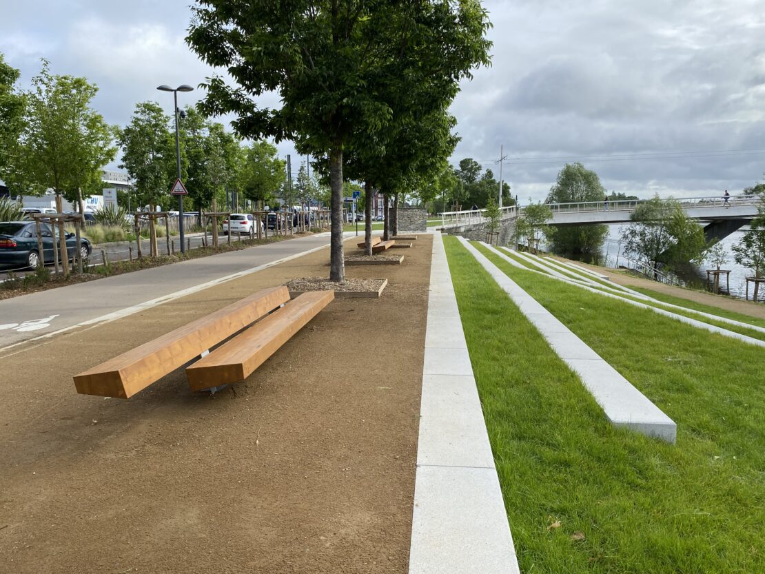 Ici, on a une vision coupée en deux : À gauche se trouvent la route, les arbres et les bancs récents, tandis qu'à droite s'étend une colline verdoyante agrémentée de murets servant de sièges.